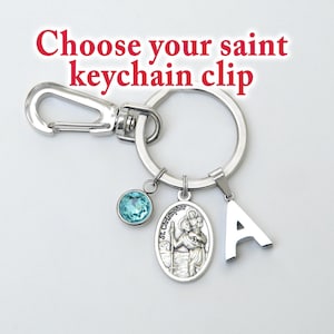 May include: A silver-toned metal keychain featuring a lobster claw clasp and a circular ring. Attached are three charms: a round light blue crystal, an oval St. Christopher medal, and the letter A. The text Choose your saint keychain clip is visible.