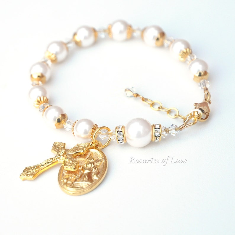 Op de afbeelding: Een witte parel en gouden rozenkrans armband met een gouden kruis en een gouden medaille met een religieus beeld. De armband heeft een karabijn sluiting. De tekst "Rosaries of Love" is zichtbaar in de afbeelding.