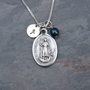 May include: A silver-toned necklace featuring an oval pendant embossed with an image of Saint Raphael and the text "S. RAPHAEL". The pendant is accompanied by a small circular charm engraved with the letter "A" and a dark blue bead charm.