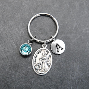 May include: A silver-toned metal keychain featuring a circular split ring with three dangling charms: a round light blue crystal, an oval-shaped medal depicting an angel with the text Guardian Angel Italy, and a small round disc with the letter A.
