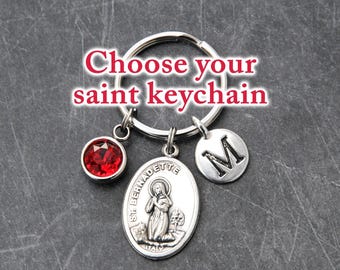 May include: A silver-toned metal keychain featuring a circular ring, a round red crystal charm, an oval medal depicting Saint Bernadette with the text 'ST. BERNADETTE ITALY', and a round charm with the letter 'M'. Text reads 'Choose your saint keychain'.