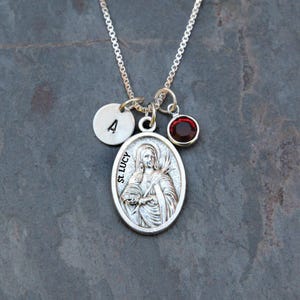 May include: A silver-tone necklace featuring an oval pendant embossed with an image of St. Lucy and the text St. Lucy. The necklace includes a small round charm stamped with the letter A and a circular charm set with a dark red gemstone.