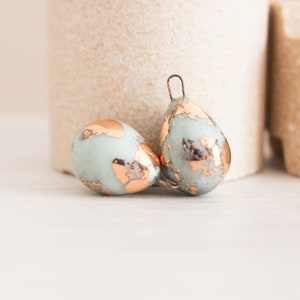 Op de afbeelding: Twee druppelvormige ornamenten met een lichtblauwe en koperen afwerking. Eén ornament ligt, de andere staat rechtop met een draadlus aan de bovenkant. De ornamenten hebben een verweerde, getextureerde uitstraling, wat wijst op een vintage of handgemaakte stijl.