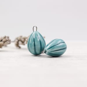 Peut inclure: Deux pendentifs en céramique bleu sarcelle en forme de goutte d'eau, avec des rayures bleu foncé et une boucle en métal pour les suspendre. Les pendentifs sont sur une surface claire, avec des brins de lavande séchée en arrière-plan.