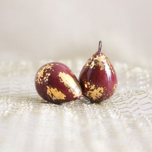 Può includere: Due ornamenti a forma di goccia con finitura bordeaux scuro e accenti in foglia d'oro. Ogni ornamento ha un piccolo anello di metallo nella parte superiore per appenderlo. Gli ornamenti sono esposti su una superficie strutturata bianco sporco.