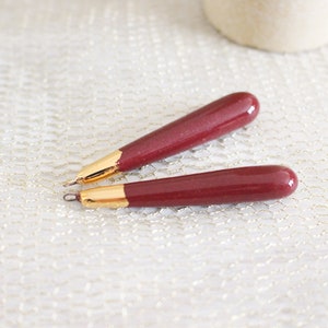 Può includere: Due penne bordeaux con punte color oro sono esposte su una superficie strutturata bianco sporco. Le penne hanno un design elegante e arrotondato, e gli accenti dorati aggiungono un tocco di eleganza. Le penne sono probabilmente usate per scrivere.