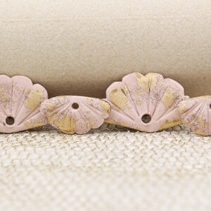 May include: Four pink seashell-shaped decorative pieces with gold accents. Each piece has a hole in the center. The seashells are arranged in a row on a textured surface.