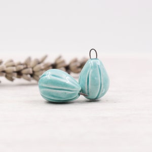 Op de afbeelding: Twee turquoise keramische druppelvormige kralen met verticale groeven. Elke kraal heeft een kleine metalen lus aan de bovenkant om te rijgen. De kralen zijn geplaatst tegen een vage achtergrond van gedroogde lavendel op een wit houten oppervlak.