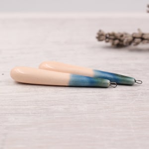 May include: Two handmade ceramic earrings. The earrings are a teardrop shape, with a light peach color at the top that fades into a blue-green hue at the bottom. Each earring has a small metal loop at the end.
