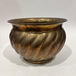 Puede incluir: Una maceta de latón vintage con un diseño en espiral. La maceta tiene una forma ancha y redondeada con un borde acampanado y una pequeña base circular. El latón tiene una pátina cálida y envejecida.