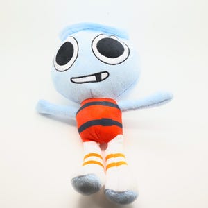 May include: A soft toy character with a light blue head and arms, large black eyes, and a smiling mouth. The torso is orange with black stripes, and the legs are white with orange stripes. The toy is designed to stand upright.
