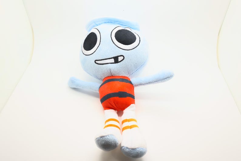 May include: A soft toy character with a light blue head and arms, large black eyes, and a smiling mouth. The torso is orange with black stripes, and the legs are white with orange stripes. The toy is designed to stand upright.