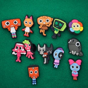 Puede incluir: Una colección de dijes de zapatos de personajes de dibujos animados coloridos. Los dijes presentan varios diseños, incluyendo un robot, un gato y otras figuras caprichosas. Están dispuestos sobre una superficie verde.