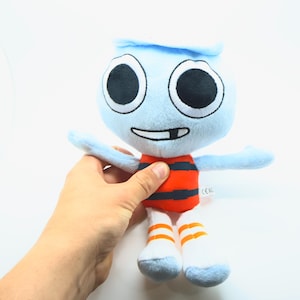 May include: A soft toy character with a light blue head and arms, large black eyes, and a smiling mouth. The toy has an orange and black striped torso, white legs, and orange striped socks. The toy is being held by a person.