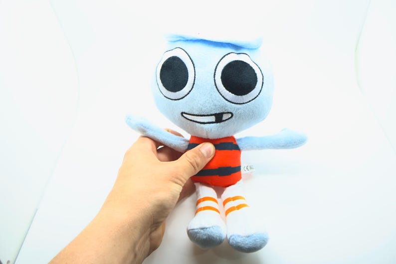 May include: A soft toy character with a light blue head and arms, large black eyes, and a smiling mouth. The toy has an orange and black striped torso, white legs, and orange striped socks. The toy is being held by a person.