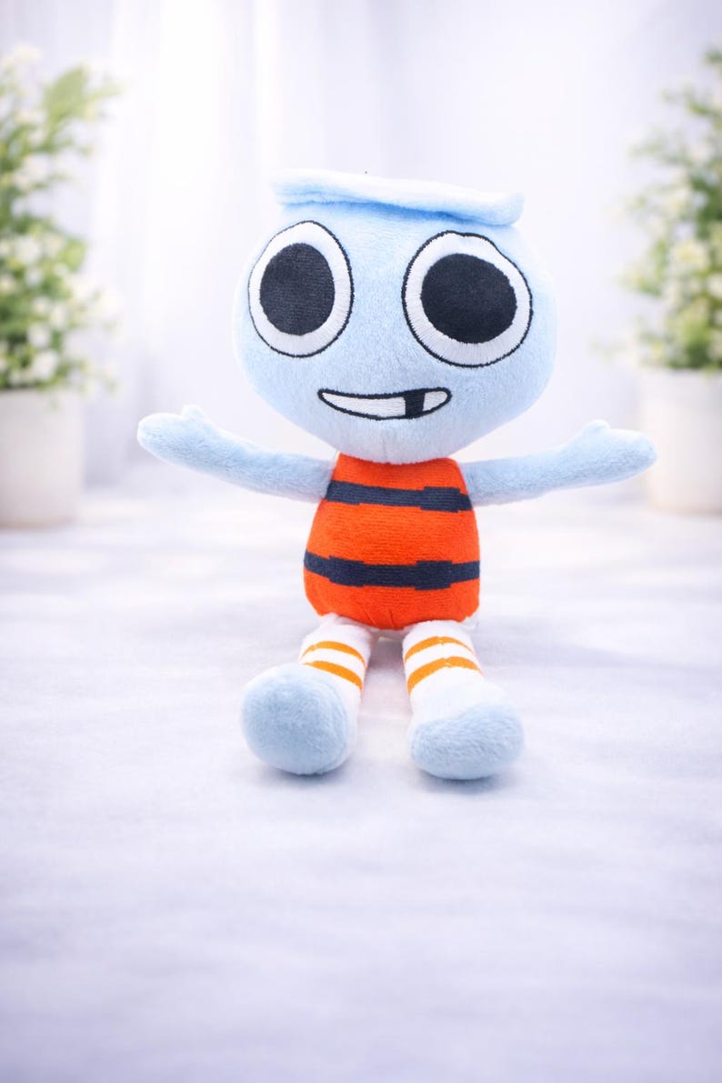 May include: A soft toy character with a light blue head and arms, large black eyes, and a smiling mouth. The body is orange with black stripes, and the legs are blue with white and orange striped socks. The toy is designed to be cuddly.