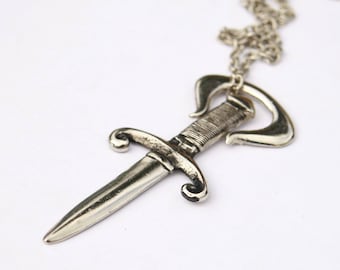 May include: A silver pendant necklace featuring a stylized dagger design. The dagger has a detailed blade and a decorative handle.