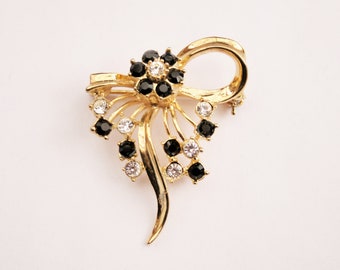 May include: A gold-toned brooch with a black and white rhinestone design. The brooch features a bow shape with a cluster of rhinestones in the center.