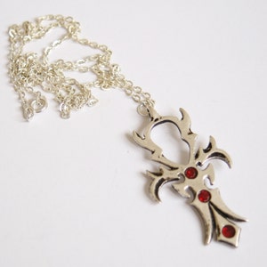 May include: Silver pendant necklace with a decorative cross design. The pendant has three red gemstones and hangs from a delicate chain. The necklace is displayed on a white background.