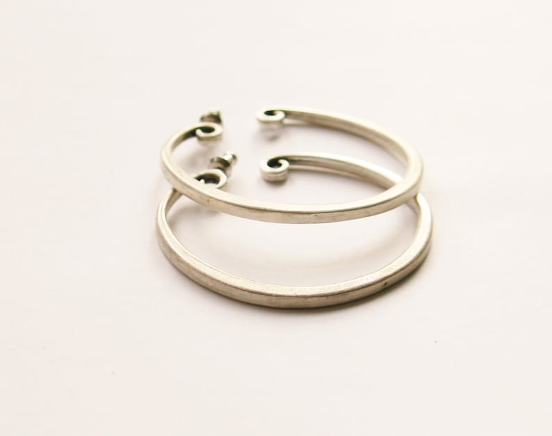 May include: A pair of silver-colored hoop earrings with a unique design. The earrings have a circular shape with a small, decorative swirl at the top. The earrings are displayed on a white background.