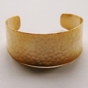 Puede incluir: Un brazalete de puño de metal martillado de color dorado. El brazalete es ancho y tiene una superficie texturizada.