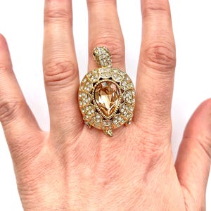 May include: A gold-tone turtle ring with a large, clear, teardrop-shaped crystal in the center. The turtle's shell is covered in smaller crystals.
