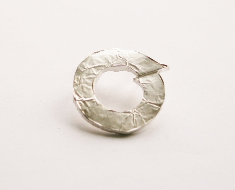 May include: A silver ring with a textured surface and a circular shape. The ring has a small opening in the center.