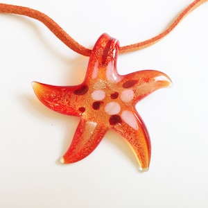 Può includere: Un ciondolo a forma di stella marina in vetro rosso e oro con dettagli bianchi appeso a un cordino in pelle marrone.