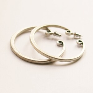 May include: A pair of silver-colored hoop earrings. The earrings have a simple, circular design with a post closure. The ends of the hoops have a decorative, curved shape. The earrings are displayed on a white background.