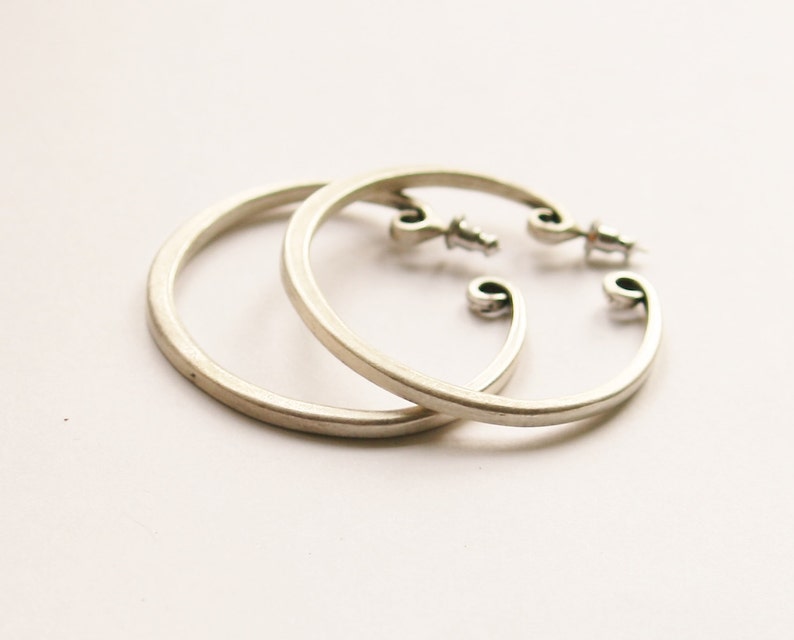 May include: A pair of silver-colored hoop earrings. The earrings have a simple, circular design with a post closure. The ends of the hoops have a decorative, curved shape. The earrings are displayed on a white background.