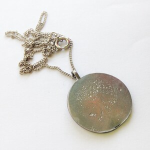 Pu&ograve; includere: Una collana a catena d'argento con un ciondolo rotondo d'argento. Il ciondolo ha una superficie testurizzata con un motivo maculato.