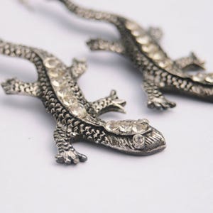 May include: A pair of silver lizard earrings with clear rhinestones. The lizards are detailed with textured scales and have dangling hooks.