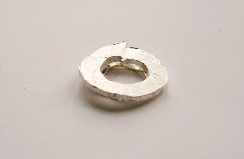 May include: A silver ring with a textured, organic shape. The ring is open and has a circular opening.