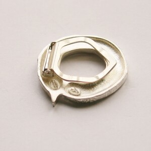 May include: Silver-toned metal belt buckle with a textured, circular design. The buckle has a hinged clasp and a small, engraved inscription.