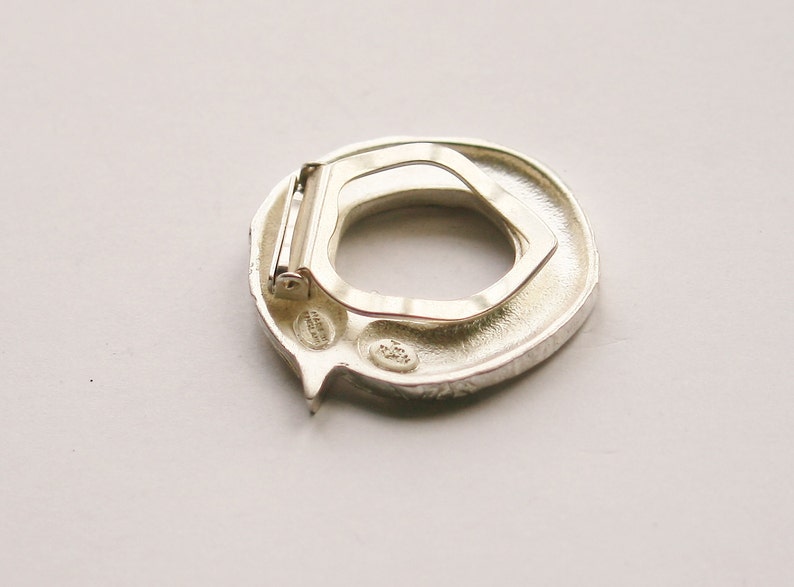 May include: Silver-toned metal belt buckle with a textured, circular design. The buckle has a hinged clasp and a small, engraved inscription.