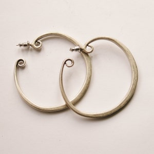 May include: A pair of silver-toned hoop earrings. Each earring has a circular design with a decorative swirl at the bottom and a post closure. The earrings are displayed on a white surface.