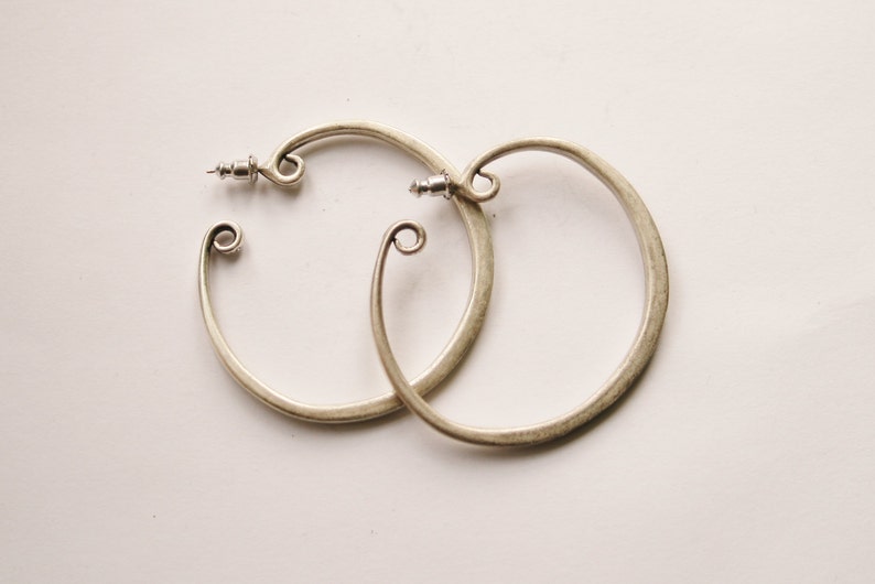 May include: A pair of silver-toned hoop earrings. Each earring has a circular design with a decorative swirl at the bottom and a post closure. The earrings are displayed on a white surface.