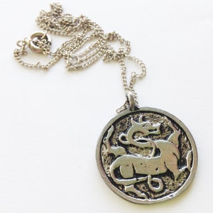 Pu&ograve; includere: Una collana con catena d'argento con un ciondolo rotondo che presenta un design di drago stilizzato e dettagliato.