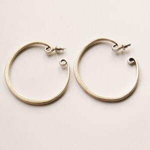 May include: A pair of silver-toned hoop earrings. Each earring has a circular design with a curled end and a post for securing. The earrings are displayed on a white surface.
