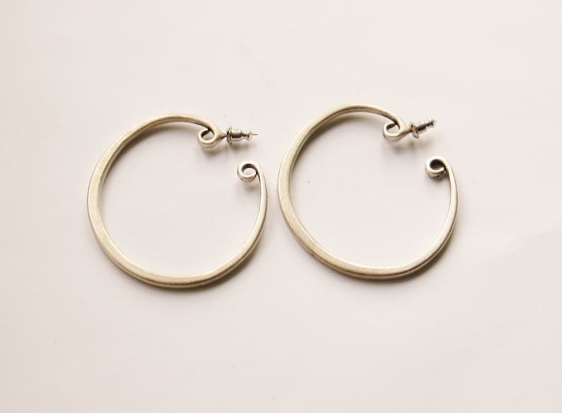 May include: A pair of silver-toned hoop earrings. Each earring has a circular design with a curled end and a post for securing. The earrings are displayed on a white surface.