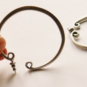 May include: Two silver-toned metal bracelets. One is held by a hand, the other lies on a white surface. Each bracelet has a circular design with decorative scrollwork at the ends and a screw closure.