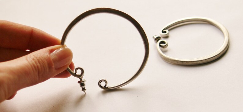 May include: Two silver-toned metal bracelets. One is held by a hand, the other lies on a white surface. Each bracelet has a circular design with decorative scrollwork at the ends and a screw closure.