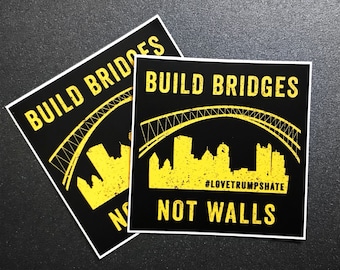 Op de afbeelding: Twee zwarte en gele stickers met de tekst "Bouw bruggen, geen muren" en een silhouet van een brug en een skyline. De kleinere sticker heeft de hashtag "#LoveTrumpsHate".