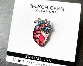 May include: A geometric enamel pin in the shape of a human heart, featuring a design of red, pink, and blue geometric shapes. The pin is on a white card with the text "1FLYCHICKEN CREATIONS" and "ENAMEL PIN".