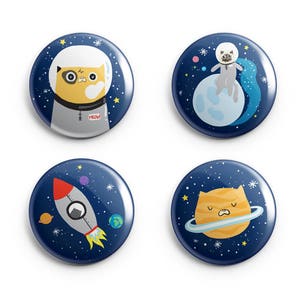 May include: Four round blue and white space-themed buttons with cat illustrations. One button features a cat in a spacesuit with the word "MEOW" on its chest. Another button features a cat sitting on a moon with stars in the background. The third button features a cat in a rocket ship. The fourth button features a cat on a planet with a ring around it.