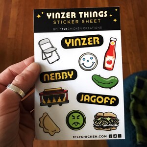 K&ouml;nnte beinhalten: Ein Aufkleberbogen mit dem Titel "Yinzer Things Sticker Sheet" von "1FlyChicken Creations". Der Bogen enth&auml;lt verschiedene Aufkleber mit Pittsburgh-Slang-Begriffen, darunter "Yinzer", "Jagoff", "Nebby" und ein Smiley mit drei Punkten. Es gibt auch einen Aufkleber einer Ketchupflasche, einer Gurke, eines Klappstuhls und eines Pierogi.
