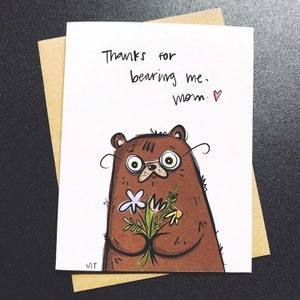 Peut inclure: Carte de v&oelig;ux avec une illustration d'ours brun portant des lunettes et tenant des fleurs. La carte porte l'inscription "Thanks for bearing me, mom." &agrave; l'encre noire, avec un petit c&oelig;ur rouge. La carte est dans une enveloppe brune.