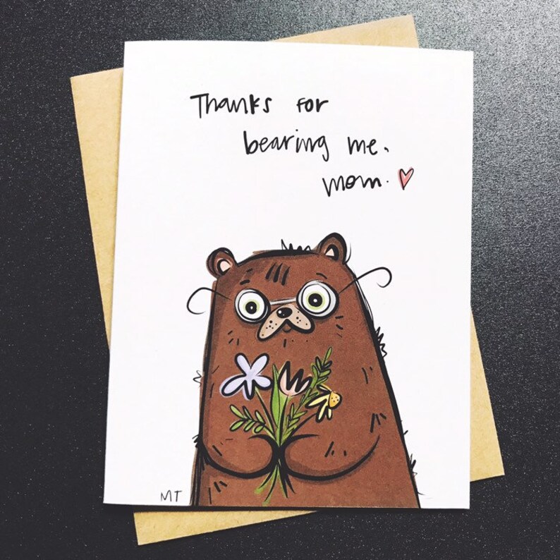 Peut inclure: Carte de v&oelig;ux avec une illustration d'ours brun portant des lunettes et tenant des fleurs. La carte porte l'inscription "Thanks for bearing me, mom." &agrave; l'encre noire, avec un petit c&oelig;ur rouge. La carte est dans une enveloppe brune.