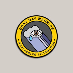 Pu&ograve; includere: Un badge circolare giallo con il testo "Gray Day Warrior Weathering Pittsburgh" attorno al bordo. All'interno del badge c'&egrave; un'illustrazione in bianco e nero di una nuvola con un occhio e una singola lacrima.