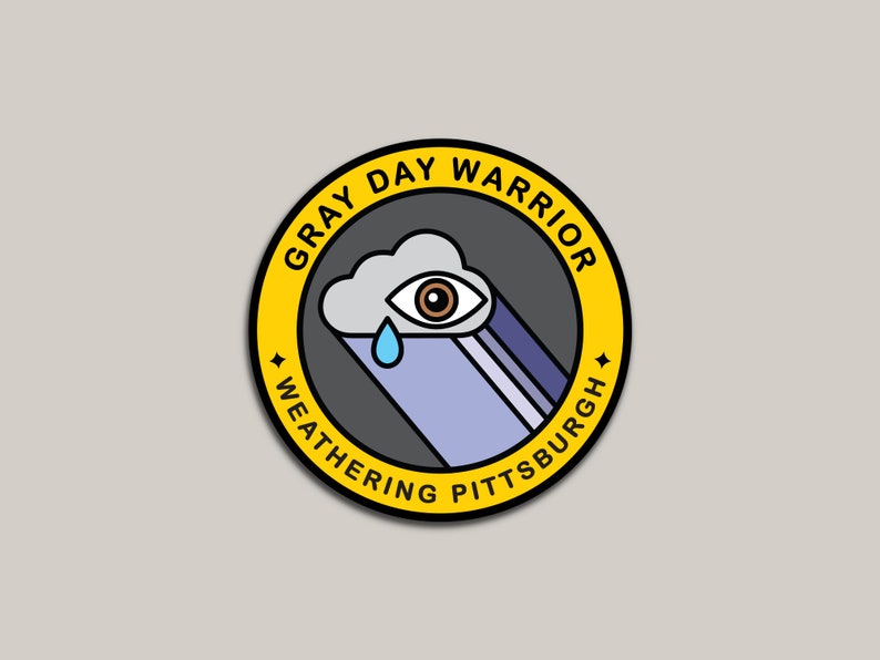 Pu&ograve; includere: Un badge circolare giallo con il testo "Gray Day Warrior Weathering Pittsburgh" attorno al bordo. All'interno del badge c'&egrave; un'illustrazione in bianco e nero di una nuvola con un occhio e una singola lacrima.
